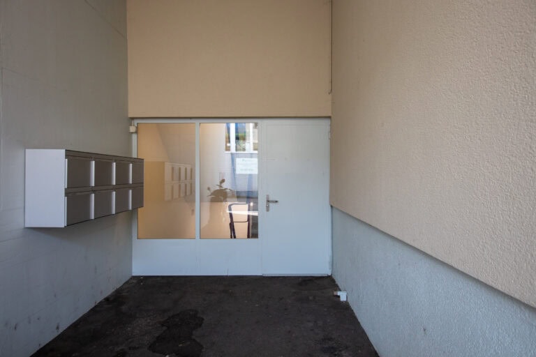 Eine Reihe von Briefkästen ist an einer Wand in der Nähe einer geschlossenen Glastür mit weißem Rahmen angebracht, wie man sie häufig in Wohnungsverwaltung oder Vermietung von Lebensräumen findet. In dem geschützten, meist schattigen Bereich sind durch die Glasscheiben Spiegelungen und eine Topfpflanze zu sehen.