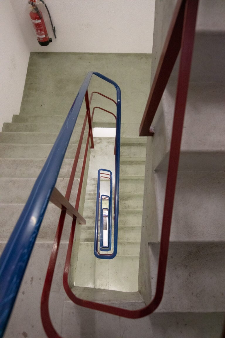Blick in ein quadratisches, mehrstöckiges Treppenhaus mit grünlich-grauen Stufen und roten und blauen Geländern - ideal für Gewerbeliegenschaften; oben links an der Wand ist ein Feuerlöscher angebracht.