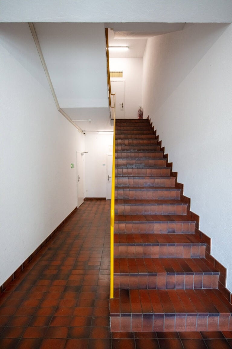 Eine Treppe mit braun gefliesten Stufen und gelbem Handlauf führt nach oben in einen weiß gemauerten Flur mit passendem braunen Fliesenboden und geschlossenen Türen auf der linken Seite - ideal für Verwaltung oder Vermietung.