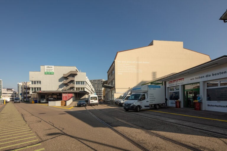 Eine Straße mit Eisenbahnschienen verläuft zwischen Industriegebäuden unter einem klaren blauen Himmel. Lieferwagen sind in der Nähe eines weißen Gebäudes mit deutschen Schildern geparkt, die auf Gewerbeliegenschaften und Vermietung hinweisen. Im Hintergrund erhebt sich ein großes beiges Gebäude.