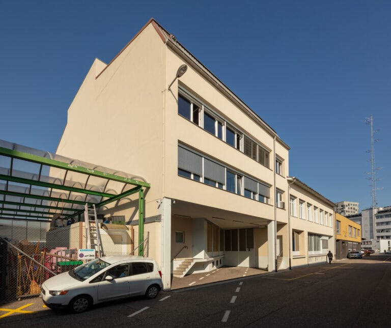 Ein dreistöckiges beigefarbenes Gebäude mit großen Fenstern steht an einer sonnigen Straße, ideal für Gewerbeliegenschaften. Ein kleines weißes Auto ist am Straßenrand geparkt, und hinter einem grün bewachsenen Zaun auf der linken Seite sind Baumaterialien zu sehen. Der Himmel ist klar und blau.