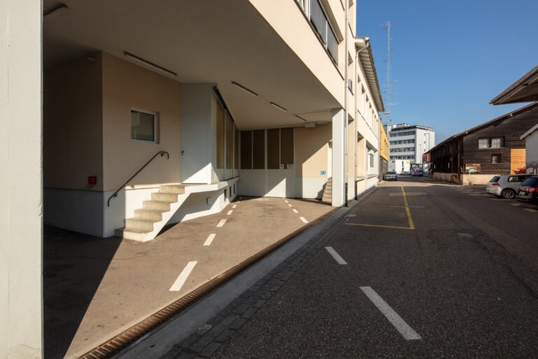 Eine ruhige städtische Straße neben modernen Gewerbeliegenschaften mit Treppen und einer Rampe, markierten Radwegen, geparkten Autos und einem klaren blauen Himmel im Hintergrund.