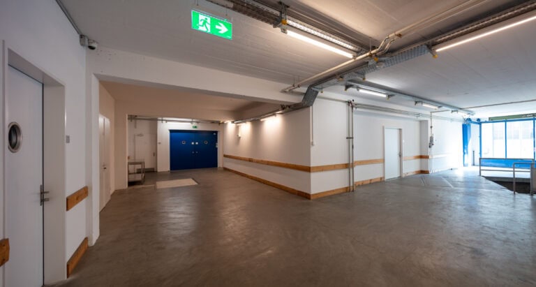 Ein sauberer, leerer Innenflur mit Betonboden, weißen Wänden, blauen Doppeltüren am Ende, einem Ausgangsschild an der Decke und Leuchtstoffröhrenbeleuchtung. Perfekt für Gewerbeliegenschaften oder neue Lebensräume. Auf der rechten Seite sind eine Rampe und eine Gittertür zu sehen.