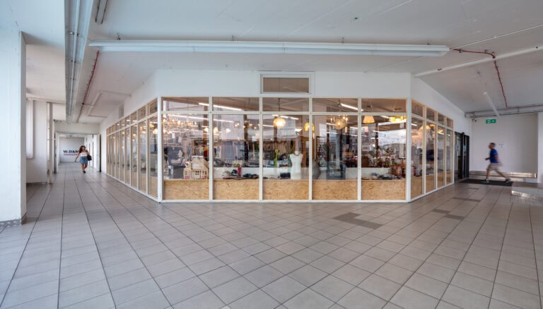 Ein gläserner Laden in einem geräumigen, gefliesten Korridor bietet verschiedene Artikel an, die typisch für Gewerbeliegenschaften sind. Zwei Personen gehen vorbei - eine in der Nähe eines Ausgangs, die andere an einem Schild - während helle Deckenleuchten diese gut geführte Verwaltung beleuchten.