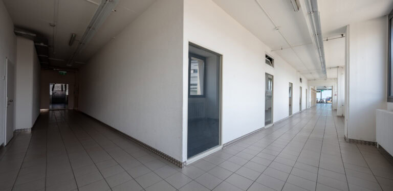 Ein heller, moderner Flur mit weißen Wänden und großen Fenstern auf der rechten Seite - ideal für Gewerbeliegenschaften - verfügt über geflieste Böden, mehrere Türen auf beiden Seiten und lässt sich in zwei Richtungen teilen, was flexible Möglichkeiten für Vermietung oder Verwaltung bietet.