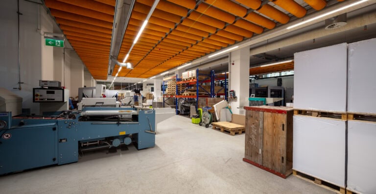 Ein moderner industrieller Arbeitsbereich mit Maschinen, großen weißen Paketen auf Paletten, Lagerregalen und einer orangefarbenen Akustikdecke. Dieser organisierte Bereich ist ein Beispiel dafür, wie Gewerbeliegenschaften für optimale Produktivität effizient verwaltet werden können.