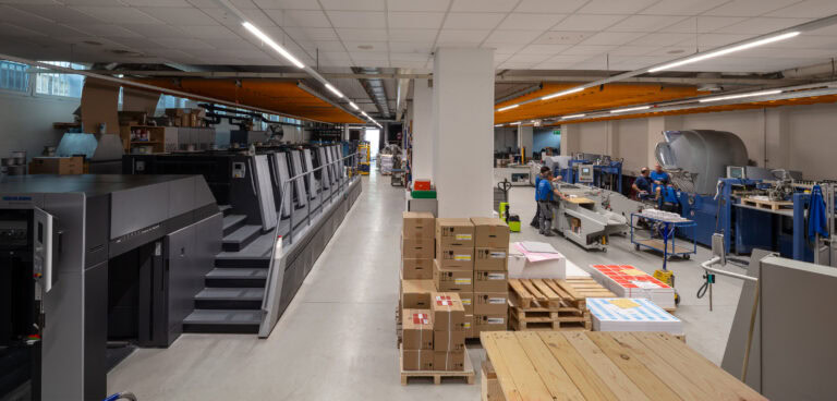 Eine geräumige, gut beleuchtete Druckerei mit großen Druckmaschinen auf der linken Seite, Stapeln von Kartons und Papier in der Mitte und mehreren Arbeitern, die Geräte bedienen, auf der rechten Seite - ideal für Vermietung und flexible Lebensräume für Ihr Unternehmen.