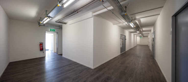 Ein sauberer, leerer Flur mit weißen Wänden, dunklen Holzböden, freiliegenden Deckenrohren und mehreren Türen. Perfekt für Verwaltung oder diverse Gewerbeliegenschaften Mietangebote, die Ausgangstür lässt natürliches Licht herein, und ein Feuerlöscher ist an der Wand montiert.