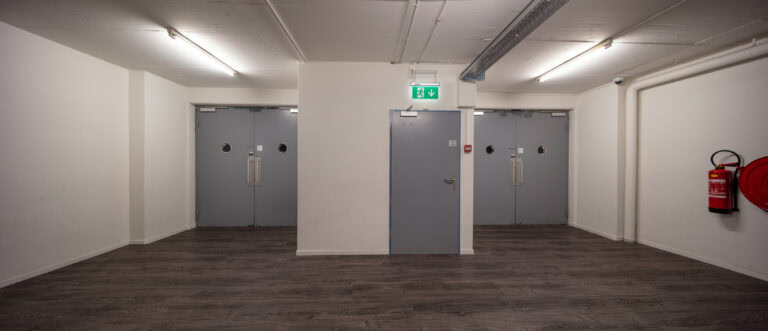 Ein Flur mit drei geschlossenen grauen Türen, weißen Wänden, holzgemustertem Bodenbelag, Oberlichtern - ideal für Gewerbeliegenschaften oder Verwaltung - mit einem Notausgangsschild über der mittleren Tür und einem roten Feuerlöscher an der rechten Wand.