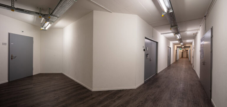 Ein sauberer, moderner Flur, ideal für Vermietung oder Verwaltung, mit weißen Wänden, grauen Türen, freiliegenden Deckenrohren und dunklem Holzfußboden. Der Korridor in diesem Wohn- oder Bürogebäude verzweigt sich in zwei Richtungen.
