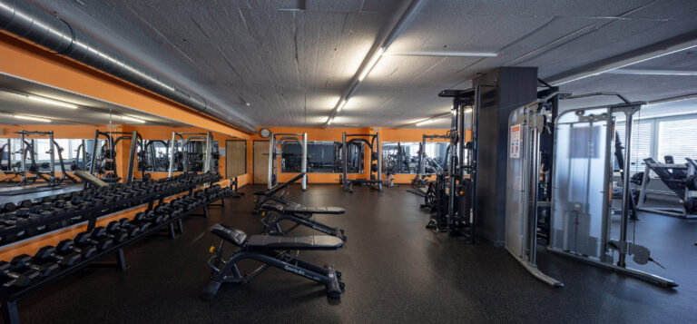 Ein geräumiges, modernes Fitnessstudio mit orangefarbenen Wänden, Hantelständern, Bänken und verschiedenen Krafttrainingsgeräten - ein ideales Mietangebot für alle, die aktive Lebensräume suchen. Große Spiegel säumen die Wände und natürliches Licht fällt durch die Fenster auf der rechten Seite ein.