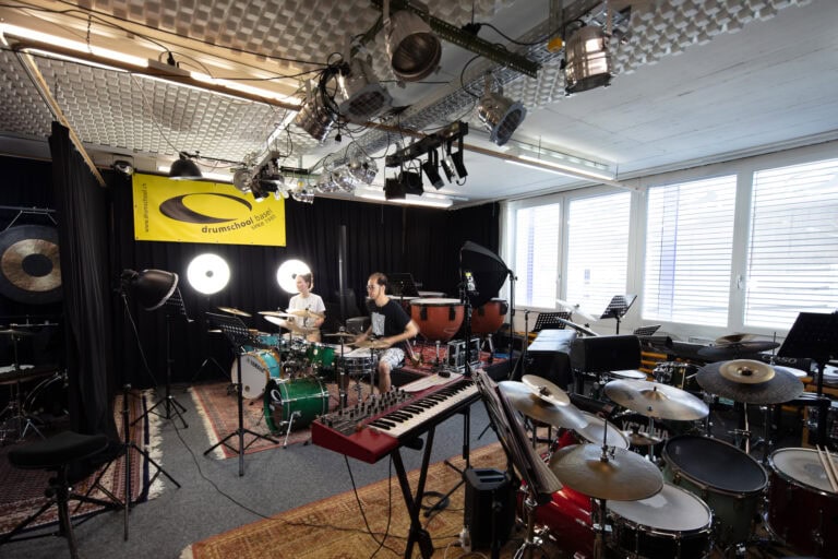 Zwei Personen spielen Schlagzeug in einem gut beleuchteten Musikstudio, das mit verschiedenen Schlaginstrumenten, Keyboards und Notenständern ausgestattet ist. Ein gelbes Schlagzeugschulbanner hängt an dem schwarzen Vorhang hinter ihnen. Große Fenster schaffen einladende Lebensräume für Kreativität.