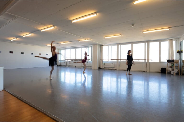 Ein geräumiges, gut beleuchtetes Tanzstudio - ideal für Gewerbeliegenschaften - mit großen Fenstern, zwei unscharfen Tänzern, die Ballettschritte üben, und einem Lehrer, der sie dabei beobachtet.