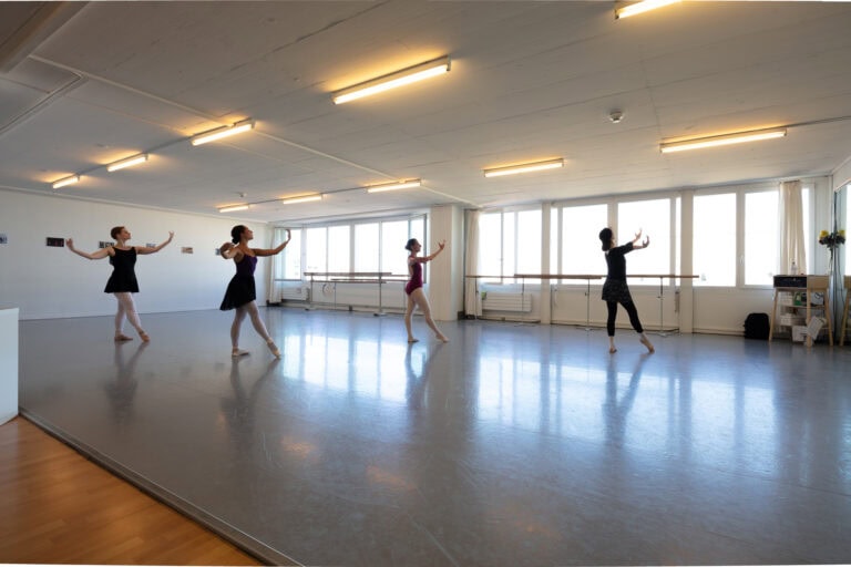 Vier Personen üben Ballett in einem geräumigen, sonnenbeschienenen Studio mit großen Fenstern und Spiegeln - ein inspirierender Lebensraum, der sich perfekt für Tanz oder Vermietung eignet, mit Holzböden am Rand und Platz für anmutige, synchronisierte Bewegungen.