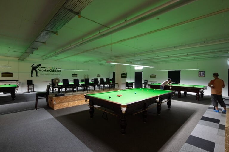 Ein geräumiger Snooker-Club mit mehreren Snooker-Tischen, zwei Spielern an einem Tisch und leeren Stühlen entlang der Wand unter grüner Deckenbeleuchtung - ideale Lebensräume zum Entspannen und Genießen.