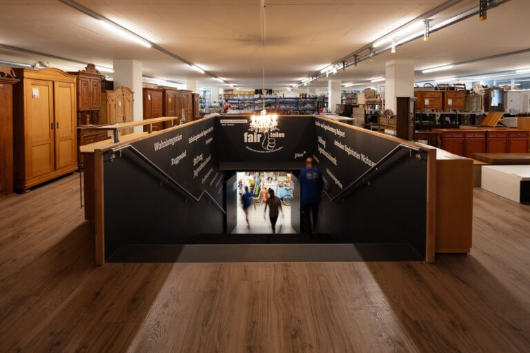 Ein geräumiges Möbelhaus mit Holzböden und verschiedenen Schränken; eine zentrale Treppe führt hinunter in eine untere Etage mit inspirierenden Lebensräumen, wo drei verschwommene Personen unter einem Kronleuchter aufsteigen.