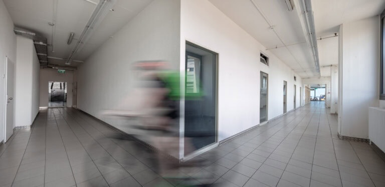 Eine unscharfe Person in Bewegung fährt auf einem Fahrrad durch einen sauberen, modernen Flur mit gefliesten Böden und weißen Wänden, was einen dynamischen Effekt in einem ansonsten leeren, gut beleuchteten Korridor mit hochwertigen Gewerbeliegenschaften erzeugt.