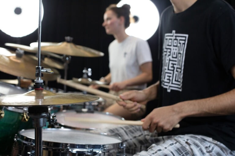 Zwei Personen spielen zusammen in einem gut beleuchteten Raum Schlagzeug. Der Fokus liegt auf den Händen und dem Schlagzeug im Vordergrund, während der andere Schlagzeuger unscharf im Hintergrund zu sehen ist - so entstehen lebendige Lebensräume für musikalische Kreativität.