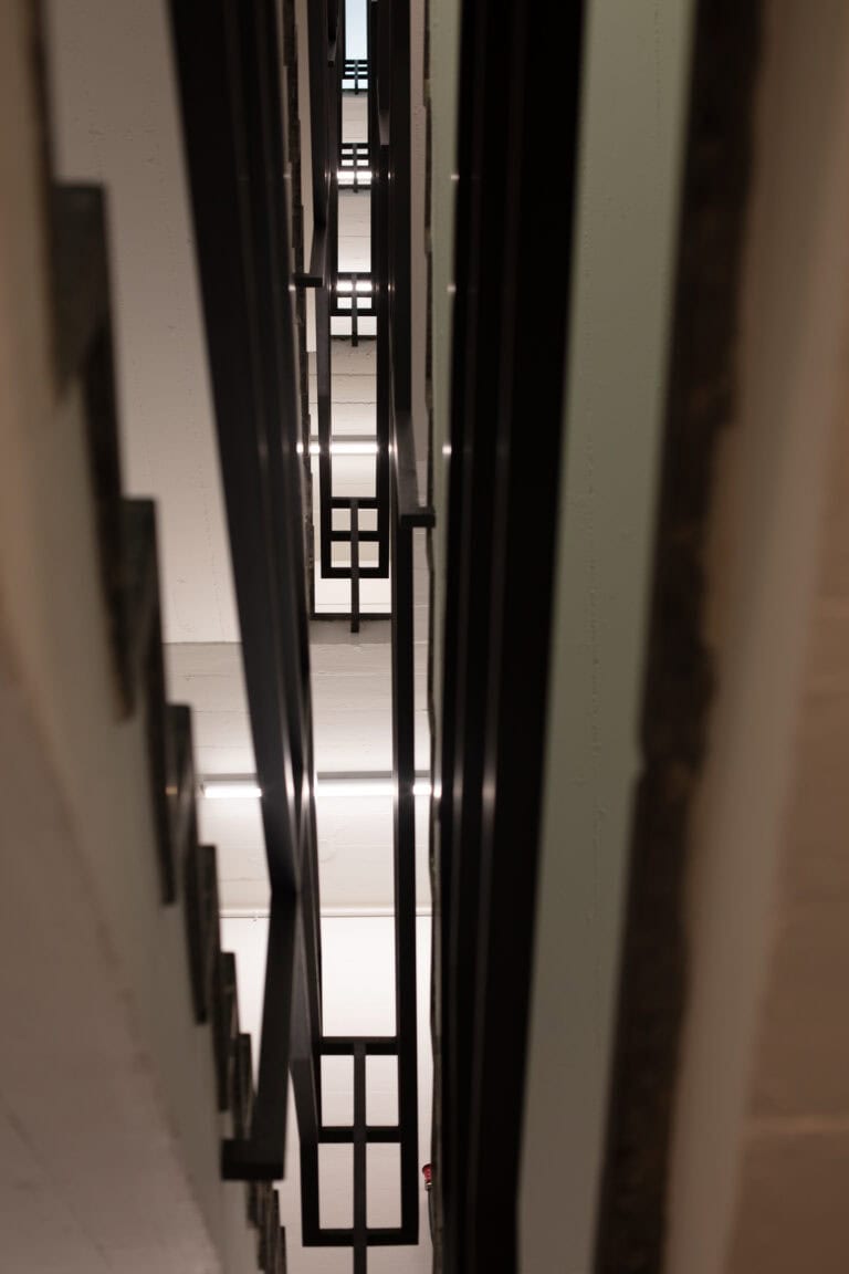 Blick auf ein schmales, hohes Treppenhaus in Gewerbeliegenschaften mit geometrischen schwarzen Geländern und weißen Wänden, die ein sich wiederholendes Muster aus Linien und Formen bilden, das mehrere Etagen hinaufführt - stilvolle Lebensräume für moderne Unternehmen.