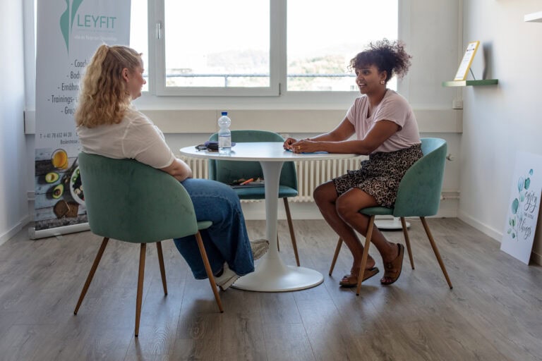 In einem hellen, modernen Büro am Leimgrubenweg 9 in Basel sitzen sich zwei Frauen an einem runden Tisch gegenüber. Die eine lächelt, während sie auf einem Klemmbrett schreibt. Auf dem Tisch steht eine Wasserflasche und im Hintergrund ist ein Fenster zu sehen.