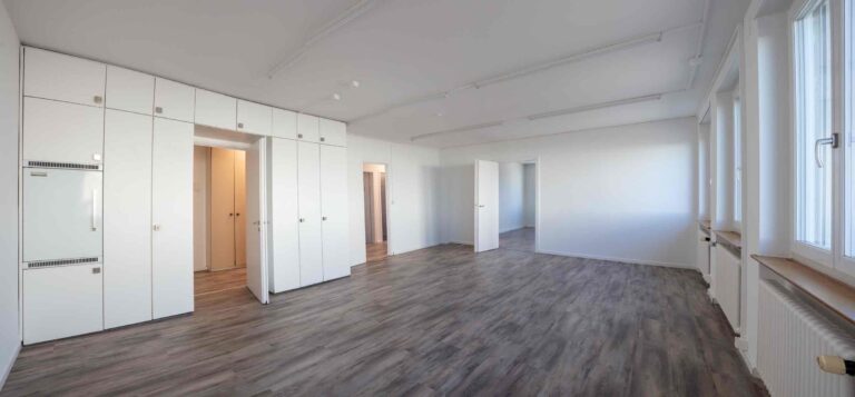 Ein heller, leerer Raum mit weißen Einbauschränken, hellgrauem Holzfußboden und großen Fenstern, die natürliches Licht hereinlassen - ideal für Gewerbeliegenschaften und Vermietung. Mehrere offene Türen führen zu angrenzenden Räumen.