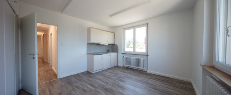 Ein kleiner, leerer Raum mit hellen Wänden und Holzfußboden, mit einer kompakten Küchenzeile, zwei großen Fenstern, einem Heizkörper und einer offenen Tür zu einem Flur - ideal für die Verwaltung oder flexible Lebensräume.