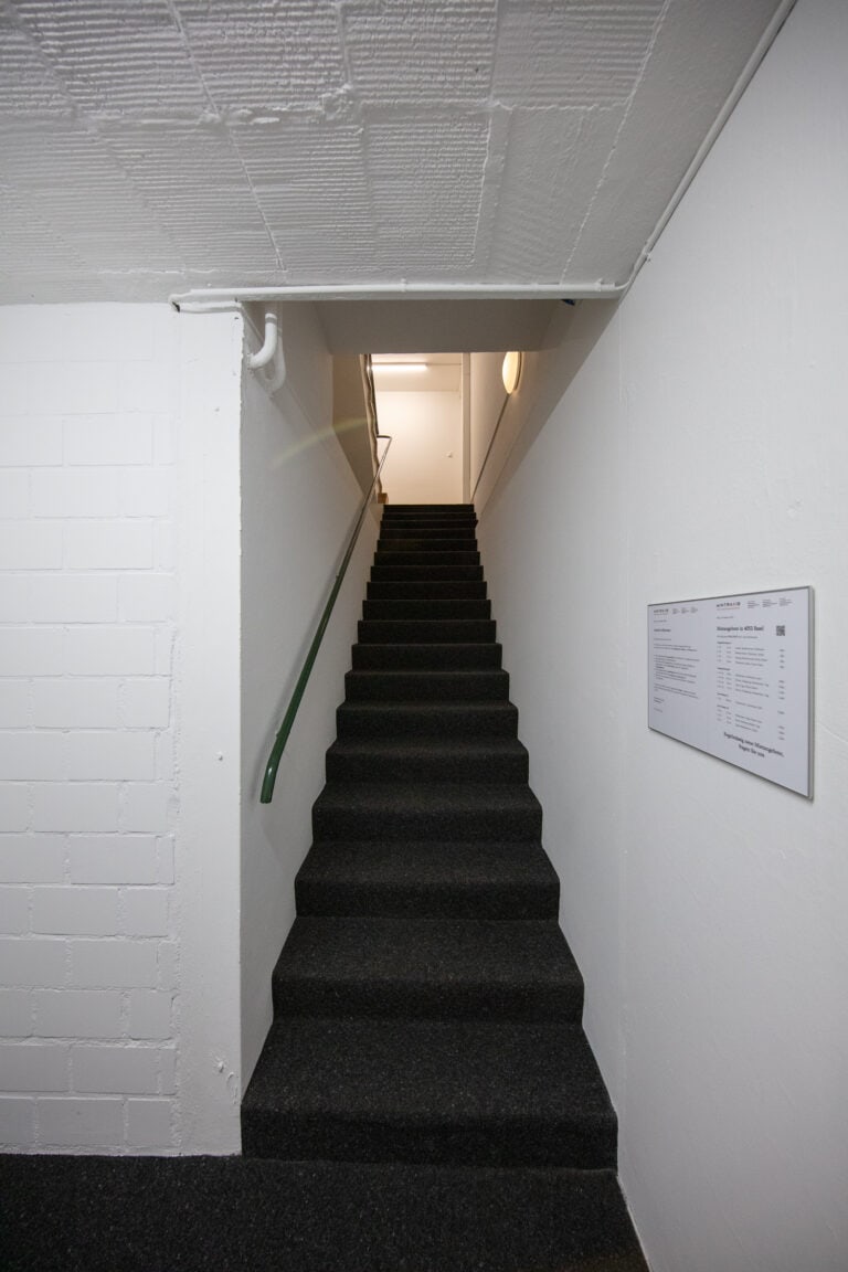 Eine schmale, mit Teppich ausgelegte Treppe mit grünem Handlauf - typisch für Gewerbeliegenschaften für Vermietung und Verwaltung - führt zwischen schlichten weißen Wänden hinauf zu einem hell erleuchteten Flur. An der rechten Wand neben der Treppe ist ein Schild mit Text angebracht.