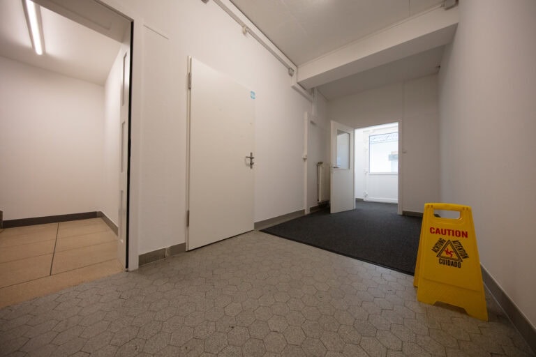 Ein Flur mit weißen Wänden und sechseckigem Fliesenboden, mit einer geschlossenen weißen Tür und einem gelben Schild 