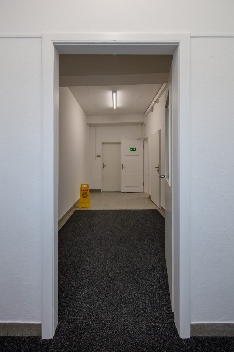 Ein Flur mit weißen Wänden und einem dunklen Teppich, der durch eine offene Tür betrachtet wird. Perfekt für Vermietung oder Verwaltung, mit einem gelben Warnschild auf dem Boden und mehreren geschlossenen Türen, von denen eine mit einem grünen Notausgangsschild gekennzeichnet ist.