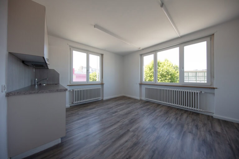 Ein heller, leerer Raum mit großen Fenstern, Holzfußboden und einem kleinen Küchenbereich erwartet die Vermietung. Natürliches Licht füllt den Raum und unterstreicht das minimalistische, moderne Design - ideal für Gewerbeliegenschaften oder flexible Nutzung.