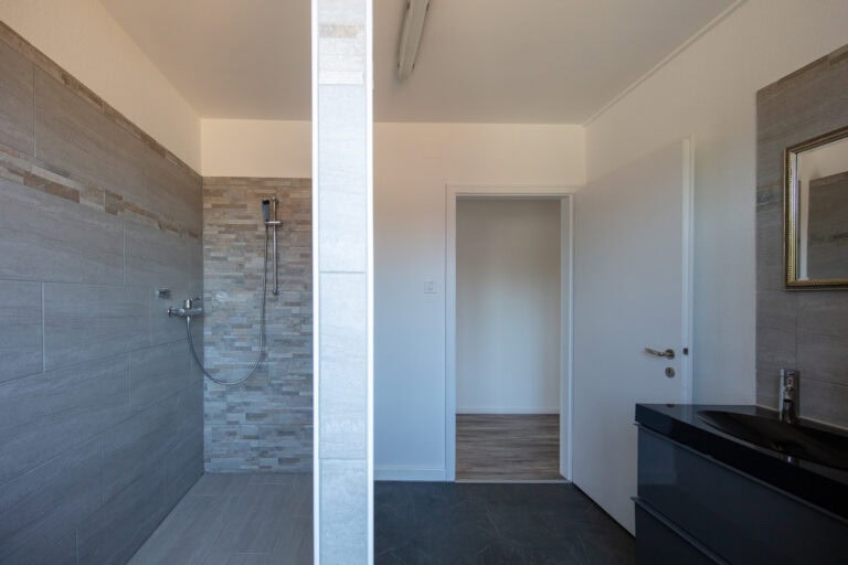 Ein modernes Bad mit grau gefliesten Wänden, einem offenen Duschbereich auf der linken Seite, einer weißen Tür in der Mitte und einem schwarzen Waschtisch mit Spiegel auf der rechten Seite - perfekt für Mietangebote oder Vermietung von stilvollen Gewerbeliegenschaften.