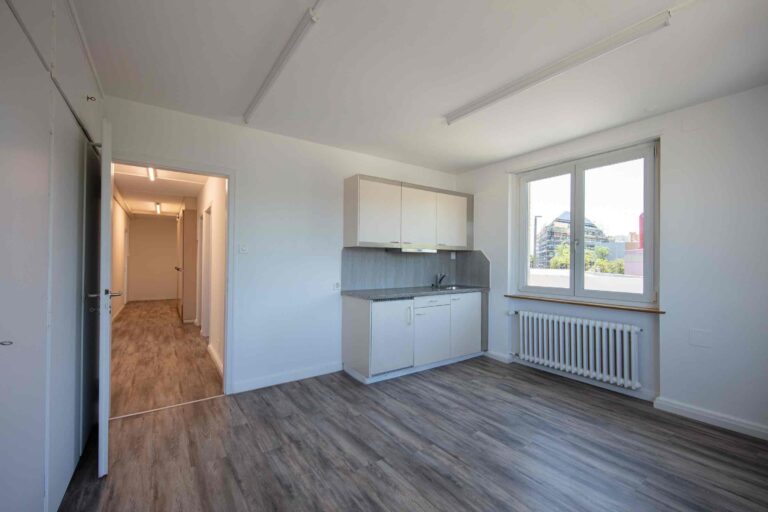 Ein helles, unmöbliertes Studio-Apartment mit Holzfußboden, weißen Wänden, einer Küchenzeile, einem großen Fenster und einem Flur zu anderen Räumen - ideale Lebensräume für alle, die Mietangebote mit viel Tageslicht suchen.