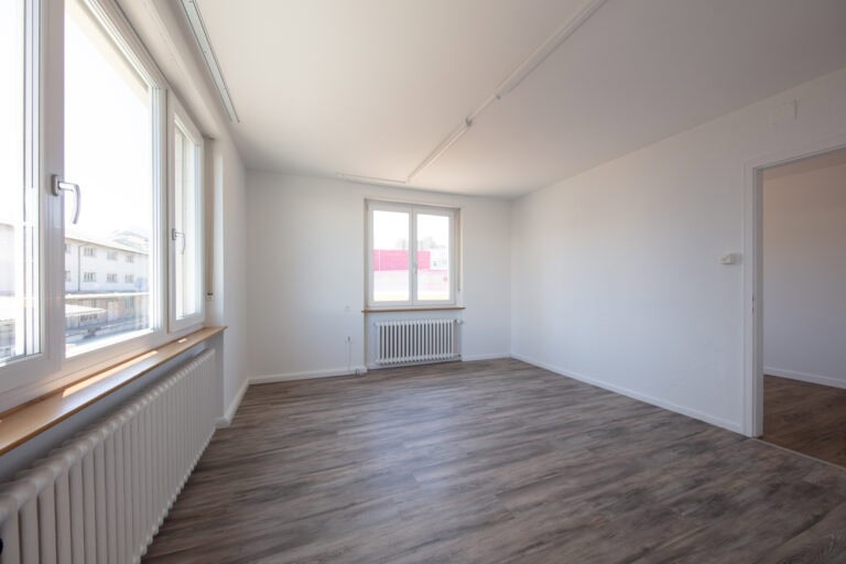 Ein leerer, heller Raum mit weißen Wänden, großen Fenstern und Holzfußböden - ideal für Gewerbeliegenschaften. Natürliches Licht erfüllt den Raum, während ein offener Durchgang zu einem anderen Raum führt. Perfekt für die Verwaltung oder diverse Mietangebote.