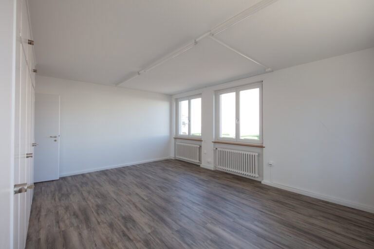Ein heller, leerer Raum mit weißen Wänden, großen Fenstern, Holzfußboden und weißen Einbauschränken auf der linken Seite. Ideal für Verwaltung oder Gewerbeliegenschaften, natürliches Licht füllt den Raum und schafft eine saubere und großzügige Atmosphäre.