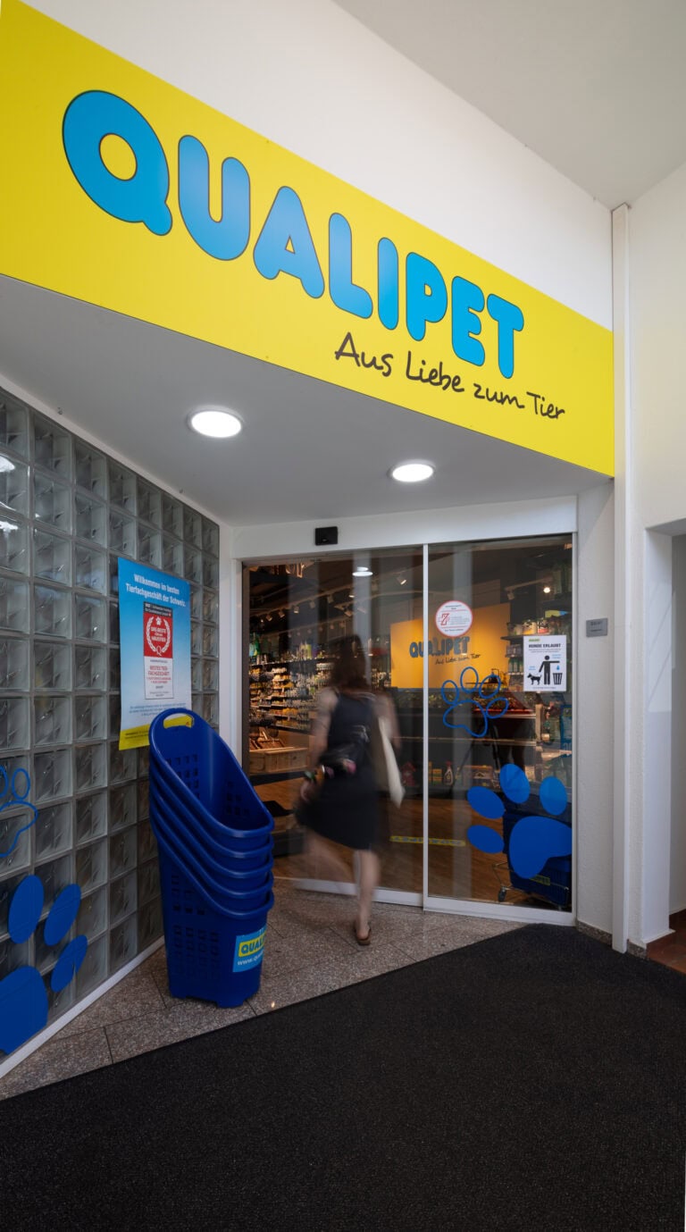 Eine Frau betritt eine QUALIPET-Tierhandlung mit einem leuchtend gelben Schild und blauen Pfotenabdrücken. Mehrere gestapelte blaue Einkaufskörbe stehen am Eingang und heißen alle tierischen Lebensräume willkommen. Über der Tür prangt der Slogan des Geschäfts 