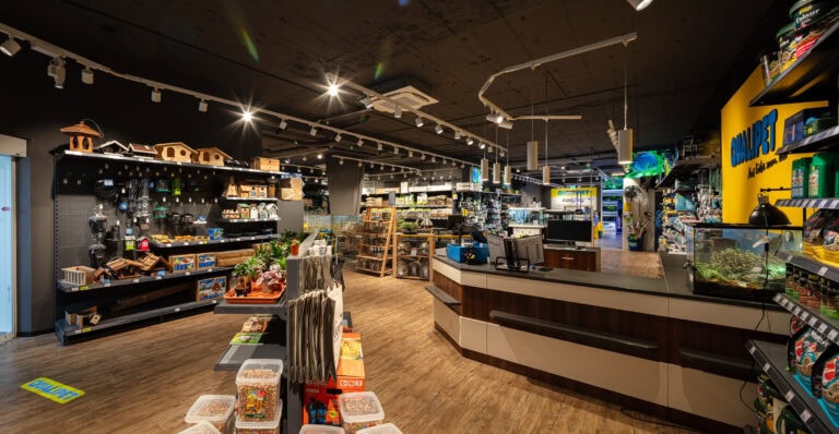 In einer hell erleuchteten Zoohandlung gibt es Regale mit Tierbedarf, Spielzeug, Vogelhäusern, Aquarien und Futter. Die Details der Verwaltung sind an der geschwungenen Kasse im Vordergrund zu sehen, und im gesamten geräumigen Geschäft sind verschiedene Produkte ausgestellt.