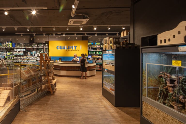 Eine Tierhandlung mit Regalen mit Produkten, Tierkäfigen und Aquarien. Zwei Personen stehen an einem Tresen mit einem gelben Hintergrund, auf dem der Name des Geschäfts steht. Der gut beleuchtete Raum mit Holzfußboden weist auch Details zu Vermietung und Mietangeboten auf.