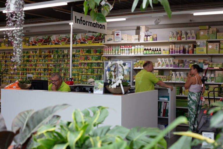 Ein Informationsschalter in einem Gartencenter mit zwei Mitarbeitern, die einer Frau helfen. Die Regale dahinter sind mit Saatgut, Gartenbedarf und Pflanzenpflegeprodukten bestückt. In der Nähe liegen auch Verwaltungsbroschüren und Mietangebote für Gewerbeliegenschaften aus. Topfpflanzen sorgen für Grün im Vordergrund.