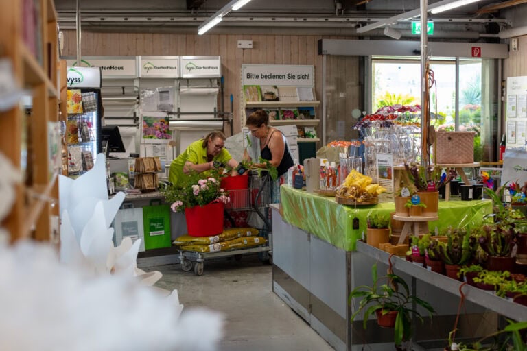 Zwei Personen arrangieren Pflanzen in einem Blumenladen. Die eine begutachtet eine Pflanze, während die andere neben einem Wagen mit Topfblumen steht. Das einladende Geschäft bietet verschiedene Pflanzen, Gartenbedarf und hebt Mietangebote für Gewerbeliegenschaften hervor.