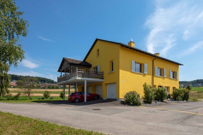 Ein knallgelbes zweistöckiges Haus mit Balkon und Garage bietet attraktive Lebensräume in einer ländlichen Umgebung mit Feldern und Bäumen. Ein rotes Auto parkt in der Einfahrt unter dem Balkon, unter einem klaren blauen Himmel.