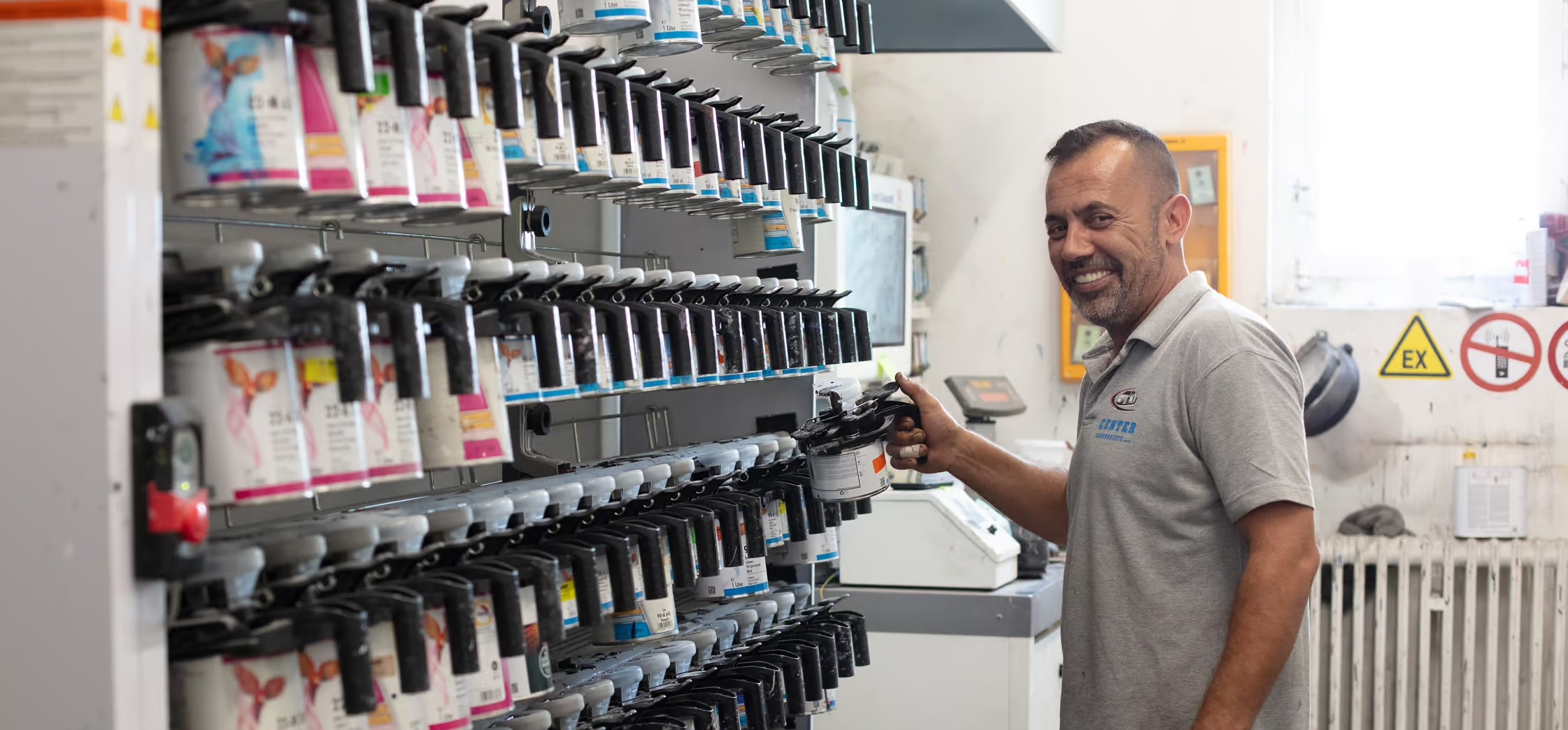 Ein Mann in einem grauen Polohemd lächelt, während er eine Farbdose in der Hand hält, die zeigt, welche Verwandlung mit den Farben der Vikara AG möglich ist. Er steht in einer Werkstatt vor Regalen, die mit verschiedenen Farbdosen gefüllt sind.