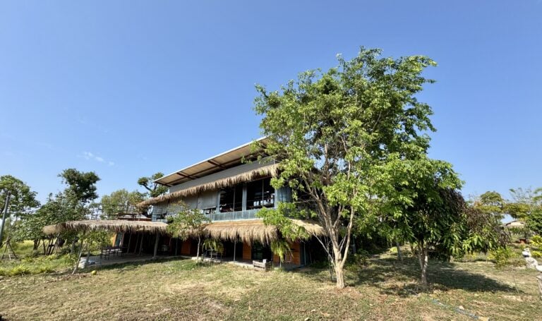 Ein zweistöckiges Gebäude mit Strohdach und großen Glasfenstern steht auf einem grasbewachsenen Grundstück, umgeben von Bäumen und Sträuchern - eine friedliche Umgebung, die unter dem klaren blauen Himmel eine Verwandlung erahnen lässt.