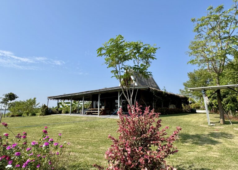 Ein rustikales Haus mit schrägem Dach steht auf einem grasbewachsenen Rasen, umgeben von Bäumen und blühenden Sträuchern unter einem klaren blauen Himmel und verkörpert den stillen Wandel der Natur im Laufe der Jahreszeiten.