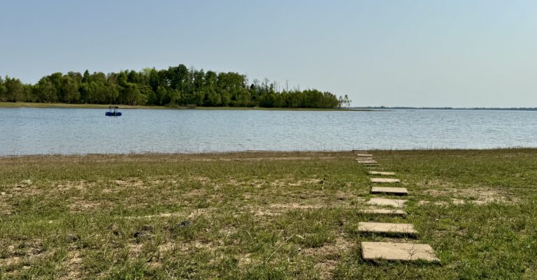 Ein steiniger Weg führt über ein grasbewachsenes, trockenes Ufer zu einem ruhigen See, an dem die stille Verwandlung der Natur offensichtlich ist - Bäume säumen das gegenüberliegende Ufer und ein kleines Boot treibt unter dem klaren blauen Himmel.