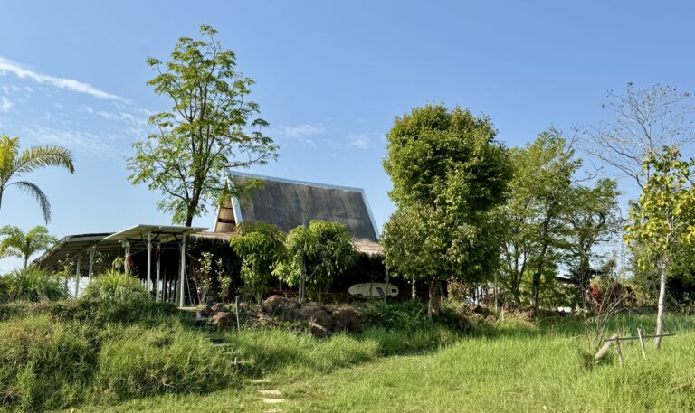 Ein kleines rustikales Haus mit steilem Dach steht inmitten üppig grüner Bäume und Gräser unter einem blauen Himmel mit Wolkenfetzen und verkörpert den Wandel. Auf der linken Seite ist eine überdachte Terrasse zu sehen, die sich nahtlos in die natürliche Umgebung einfügt.