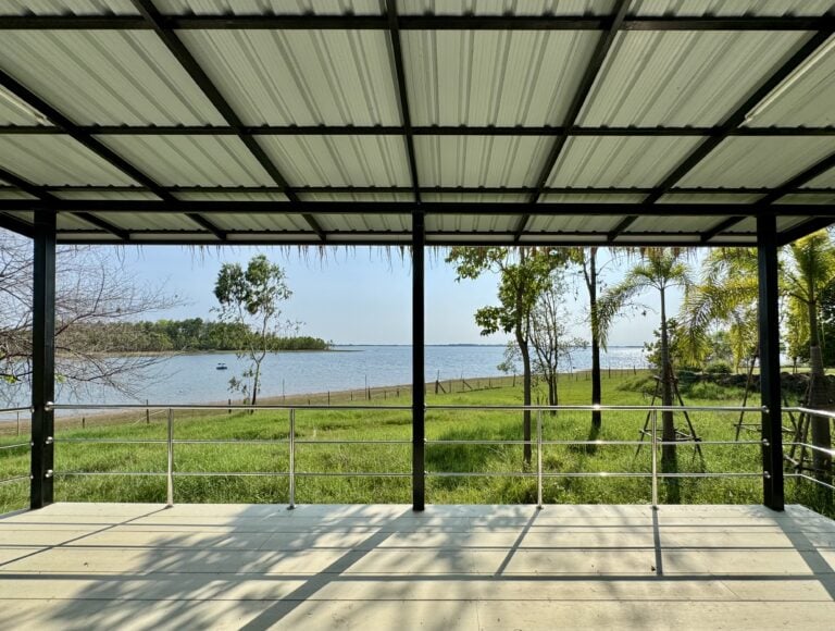 Blick von einer überdachten, offenen Terrasse mit Metallgeländer auf grünes Gras, Bäume und einen ruhigen See unter strahlend blauem Himmel - eine ruhige Umgebung, die zum Nachdenken und zur Transformation einlädt.