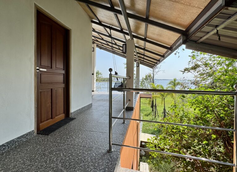 Ein überdachter Außenkorridor mit Holztür, Metallgeländer und grauem Kachelboden zeigt die Verwandlung des Raums und bietet einen Blick auf einen grünen Garten mit Bäumen und einen weiten Blick auf das Wasser unter einem klaren Himmel.