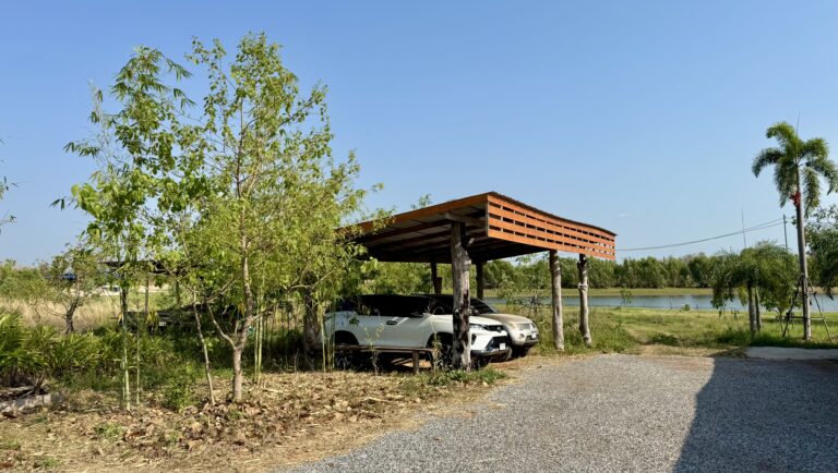 Zwei Autos sind unter einem kleinen hölzernen Carport zwischen Bäumen und einer Kiesauffahrt in der Nähe eines Teiches geparkt - eine friedliche Szene, die den Übergang von der ungezähmten Natur zu einer gepflegten Landschaft unter dem klaren blauen Himmel widerspiegelt.