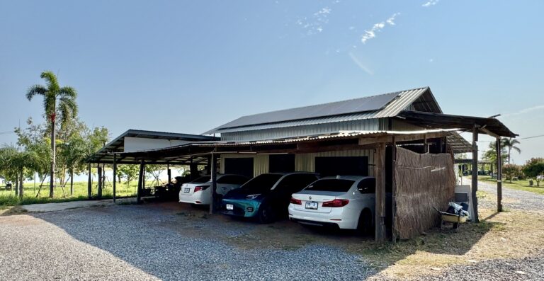 Ein metallüberdachter Carport mit vier geparkten Autos auf einem Kiesplatz steht neben einem kleinen Gebäude, das die Umwandlung des Raums veranschaulicht. Im Hintergrund sind Palmen und Gras unter einem klaren, blauen Himmel zu sehen.